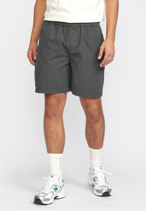 Grijze katoenen shorts met een elastische tailleband, zijzakken en een gestikte zoom, gedragen met witte en groene sport schoenen en lichte sokken.