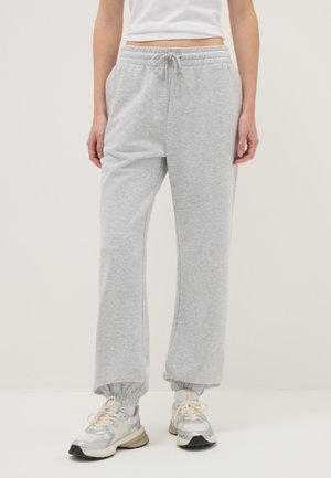 Personne portant un pantalon de survêtement gris clair à cordon de serrage avec des poignets élastiques et des baskets blanches, debout devant un fond uni.