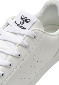 Hvit lær sneakers med perforert detaljer, flate hvite lisser og et logomerke med en mørk design på tungen.