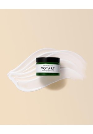 Pot en verre vert avec un couvercle noir, étiqueté "Crème Nutriente Légère Votary." Entouré d'un tourbillon de crème lisse de couleur blanche sur un fond beige.