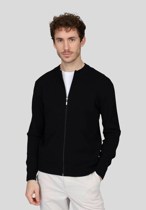 Un pull zippé noir avec un col rond, des manches longues et des poignets côtelés. Porté sur un t-shirt blanc, le tout sur un fond gris.