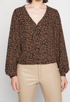 Woman wearing a black V-neck blouse with small orange floral print and beige pants, shown from shoulders to hips.