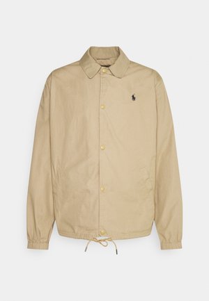 Veste beige à boutons avec col, poignets élastiques, cordon de serrage à l'ourlet, poches latérales et petit logo brodé foncé sur le côté gauche de la poitrine.
