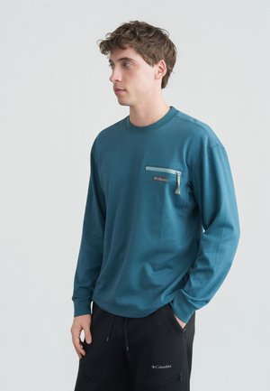 Joven con camiseta de manga larga color verde azulado con bolsillo con cremallera en el pecho y pantalones negros, con las manos parcialmente en los bolsillos, de pie contra un fondo blanco.