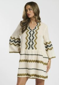 Robe couleur crème avec des motifs en zigzag en vert olive et noir, présentant un décolleté en V, des manches évasées et une jupe à volants.