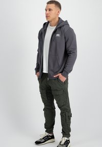 Felpa grigia con zip e tasche, indossata sopra una maglietta bianca, abbinata a pantaloni cargo verdi e scarpe da ginnastica nere. Stile casual, vestibilità rilassata.