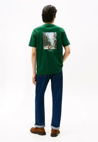 Grøn bomulds T-shirt med et trykt bybillede p�å ryggen. Den har rund hals og korte ærmer, som er parret med blå jeans og brune sko.