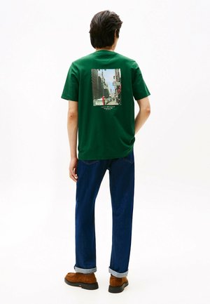 Grøn bomulds T-shirt med et trykt bybillede på ryggen. Den har rund hals og korte ærmer, som er parret med blå jeans og brune sko.