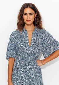 Robe à motifs paisley bleus et blancs avec des manches courtes, un décolleté en V et un détail de taille cintrée. Tissu lisse et fermeture à bouton visible.