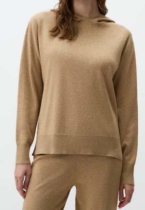 Femme portant un haut en maille beige à manches longues et un pantalon en maille assorti, debout devant un fond clair uni.