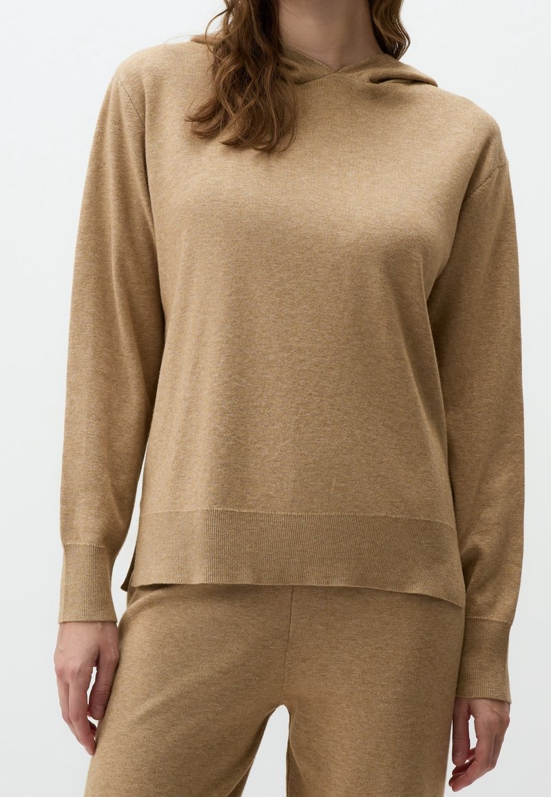 Femme portant un haut en maille beige à manches longues et un pantalon en maille assorti, debout devant un fond clair uni.