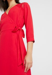 Robe portefeuille rouge avec des manches trois-quarts, des détails en dentelle sur l'épaule et une ceinture nouée à la taille. Tissu lisse avec une silhouette structurée.