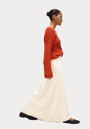 Kvinde gående i en tekstureret orange sweater, lang hvid nederdel, sorte flade sko, med håret i en knold og guld øreringe.