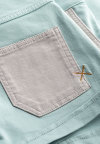 Tissu bleu clair avec une poche rectangulaire grise présentant des coutures visibles et un détail de broderie croisée orange. Texture lisse.