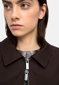 Brązowa kurtka z wyraźnym kołnierzem, z zamkiem błyskawicznym i błyszczącymi akcentami przy dekolcie. Gładka faktura z minimalnymi detalami.