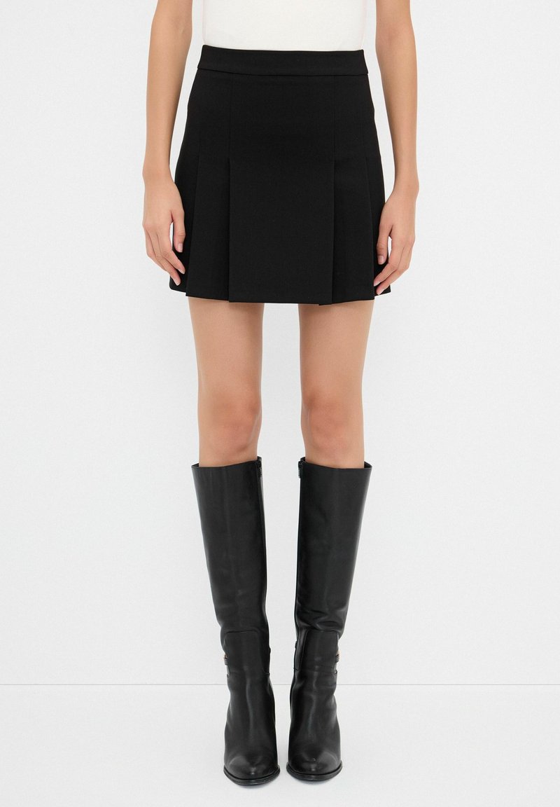 Jupe mini plissée noire avec une taille ajustée, associée à des bottes en cuir noires montantes dotées de fermetures éclair et d'embouts pointus.