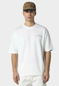 Hvid bomulds T-shirt med korte ærmer, med "PAS DÉ MONACO" i guld broderi på brystet. Sammen med en brun kasket.