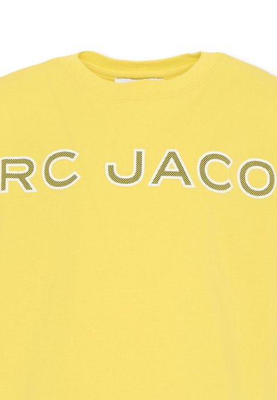 Camiseta amarilla de cuello redondo con texto parcial en negrita "RC JACO" en un patrón de cuadros blancos y negros en el pecho.