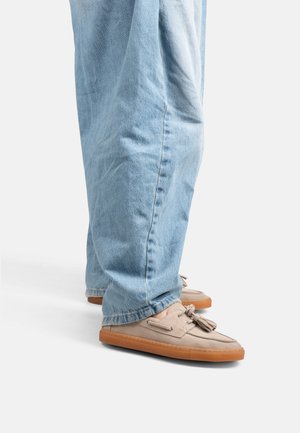 Pantalones vaqueros azul claro combinados con mocasines beige de ante con borlas y suelas de goma en un fondo blanco.