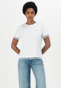 Camiseta de algodón blanca de manga corta, con un ribete plateado y un patrón sutil en los puños y el dobladillo. Detalle del logo dorado en el pecho.