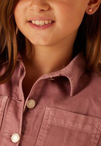 Roze knoopshirt met kraag, voorzien van een borstzak en metalen knopen. Close-up van een kind dat een hartvormige oorbel draagt.