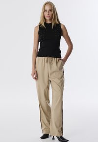 Top nero senza maniche con borchie argentate e design aderente, abbinato a pantaloni beige a gamba larga con vita regolabile. Scarpe col tacco nere.