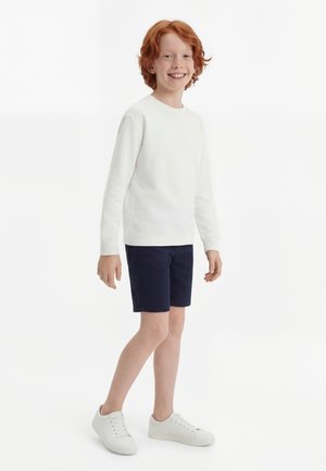 Naeratav punase juuksevärviga laps, kes kannab valget pikkade varrukatega särk, tumesiniseid lühikuid ja valgeid tossusid, seisab valge tausta ees.