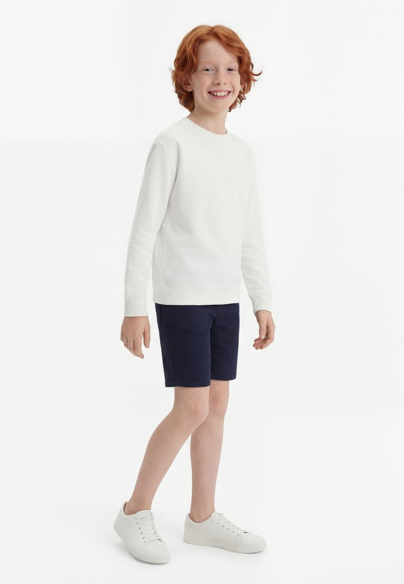 Χαμογελαστό παιδί με κόκκινα μαλλιά που φοράει λευκό πουκάμισο μακρυμάνικο, μπλε σορτς και λευκά αθλητικά παπούτσια, στέκεται μπροστά σε λευκό φόντο.