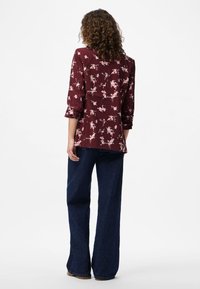 Kvinne med krøllete hår iført en burgunderblomstret bluse og vide blå jeans, stående mot en ensfarget hvit bakgrunn, med ryggen mot oss.