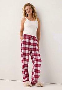 Pantaloni del pigiama a quadri rossi e bianchi con elastico in vita e tasche, abbinati a una canottiera bianca e ciabatte beige soffici.