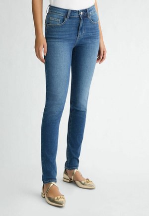 Kobieta w średnio niebieskich wąskich dżinsach z wysokim stanem i złotych, spiczastych balerinach z krzyżowym paskiem, stojąca na prostym białym tle.