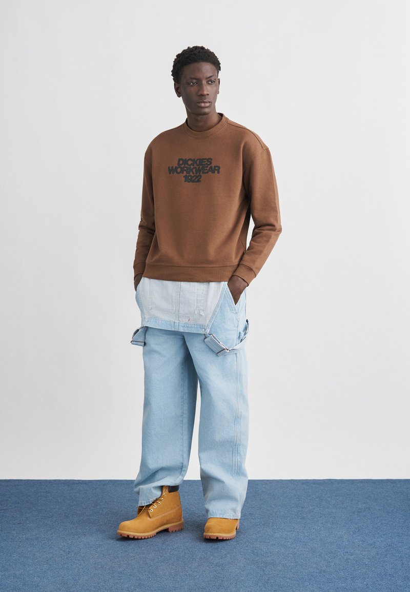 Man wearing brown sweatshirt with "Dickies Workwear 1922," light blue baggy jeans with apron-style detail, and tan work boots on blue floor.