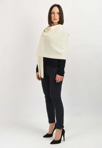 Beige uld tørklæde draperet over skuldrene, blød tekstur, vist sammen med en sort tætsiddende top og mørke jeans. Sorte spidse højhælede sko fuldender outfittet.