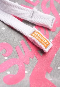 Tessuto grigio con grande testo rosa glitterato e pois bianchi; include una tracolla bianca con un'etichetta arancione Superdry.