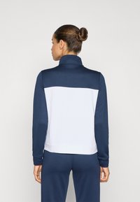 Casaco desportivo com fecho zip em azul-marinho e branco, apresentando um colarinho alto, mangas longas, tecido suave e um design em blocos de cor na parte de trás.