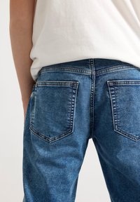 Blue denim jeans with a faded texture, featuring two back pockets and contrast stitching, paired with a simple white T-shirt.