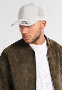 Heled hall snapback müts, millel on võrgupaneelid, kõver nokk ja bränditud plaat. Kandmine oli oliivrohelise pommikotiga ja valge täpilise särgiga.