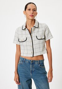 Kortærmet jakke i hvid og sort tweed, med to frontlommer, sort piping og guldknapper. Båret med blå jeans.