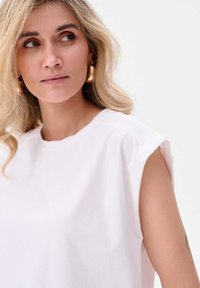 Witte katoenen mouwloze top met een ronde halslijn, voorzien van subtiele schouderdetails. Het model heeft lang golvend blond haar en draagt gouden oorbellen.