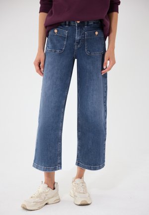 Personne portant un jean bleu à jambes larges et raccourcies avec des poches plaquées à l'avant et des baskets beige, debout devant un fond uni.