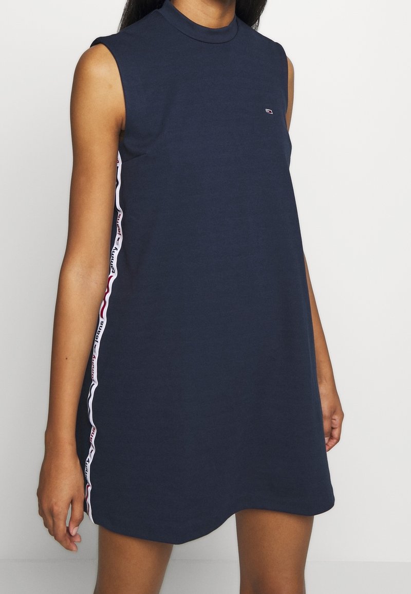 Navy sininen hihaton mekko, jossa on korkea kaula-aukko ja sivurei'ät, featuring valkoisia ja punaisia brändättyjä raitoja sivuilla. Valmistettu pehmeästä kankaasta.