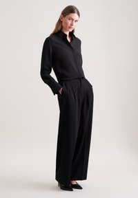 Camicia nera con bottoni e maniche lunghe, abbinata a pantaloni neri a vita alta e gamba ampia. Tessuto morbido, design minimalista, décolleté nere a punta.