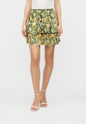 Nainen, joka pitää päällään kerroksellisen kukkakuosisen minihameen, jossa on vihreitä lehtiä ja keltaisia kukkia, yhdistettynä valkoisiin nilkkaremmiin ja korkoihin.