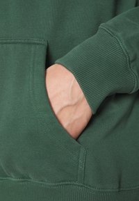 Handen inskjuten i framfickan på en grön sweatshirt med ribbat ärmslut.