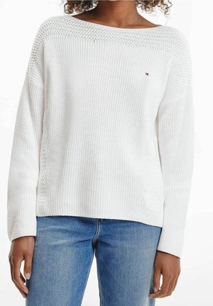 Frau trägt einen weißen Strickpullover mit U-Boot-Ausschnitt und langen Ärmeln, kombiniert mit blauen Jeans, knapp über der Taille abgeschnitten.