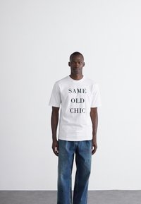 White cotton t-shirt with black text reading "SAME OLD CHIC," paired with blue oversized denim jeans. Simple, casual design.