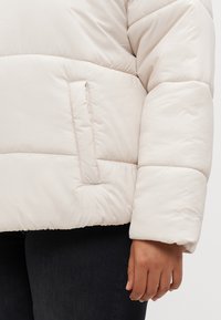 Φουσκωτό, ανοιχτό μπεζ μπουφάν με καπιτονέ σχέδιο, που διαθέτει μια πλάγια τσέπη και ανάγλυφο λογότυπο στο μανίκι, κατασκευασμένο από μαλακό, επενδεδυμένο ύφασμα.