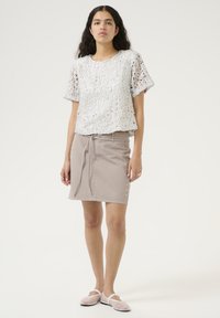 Weißes Häkeloberteil mit kurzen Ärmeln, kombiniert mit einem beigen Rock mit Gürtel und seitlichen Knöpfen, weicher Textur und flachen Schuhen.