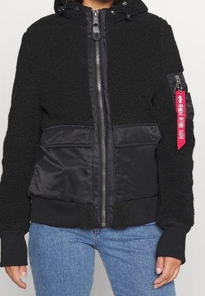 Czarna bluza zapinana na suwak z polaru z kapturem, dwie przednie kieszenie z patkami, ściągacze na rękawach oraz czerwona metka "Remove Before Flight" na rękawie.