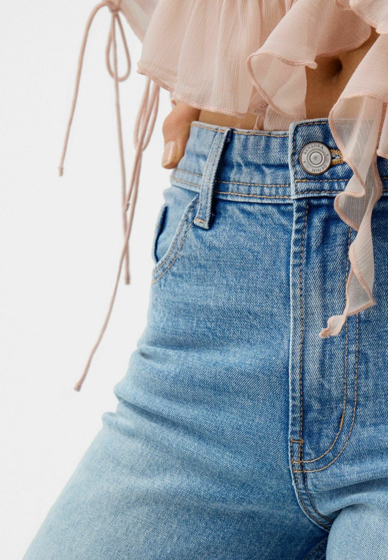 Hellblaue, hoch geschnittene Jeans mit einer schmalen Silhouette, fünf Taschen und einem Metallknopfverschluss. Kombiniert mit einem sanft rosa, rüschengeschmückten Oberteil.